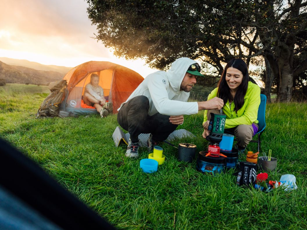 Two people prepare a meal with a backpacking stove as the sun sets behind a woman in a tent.